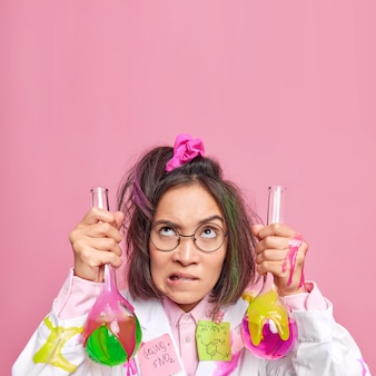 Wissenschaftliche mitarbeiterin beißt lippen hält proben chemischer flüssigkeit, die nach oben konzentriert ist und mit der forschung beschäftigt ist, trägt brille und weißen kittel auf rosafarbenem leerem kopienraum