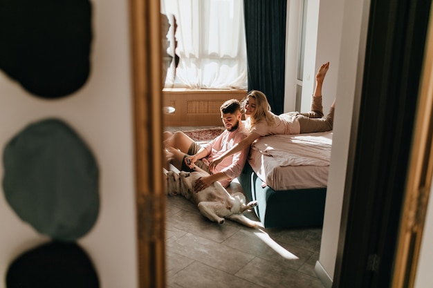 Wochenendfoto des Paares, das mit Labrador im sonnigen Schlafzimmer spielt. Kerl und Mädchen streicheln Bauch des großen Hundes.