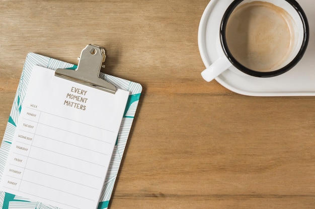 Wöchentlicher Plan auf Klemmbrett und Tasse Kaffee auf hölzernem Schreibtisch