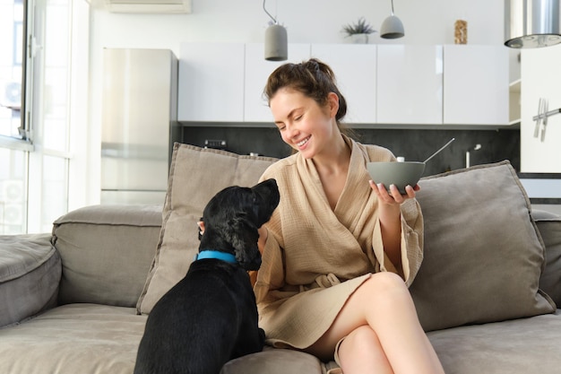 Wohn- und Lifestyle-Konzept schöne junge Frau mit Hund sitzt auf dem Sofa, isst Müsli und umarmt sich