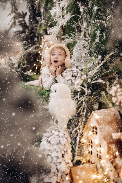 Wonderes kaukasisches Kind mit langen blonden Haaren liegt in Weihnachtsatmosphäre mit vielen geschmückten Bäumen um sie und Eule