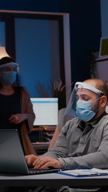 Workaholic-Geschäftsmann mit Gesichtsmaske und Visier gegen Covid, das im Firmenbüro arbeitet