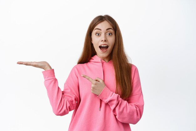 Wow, schau hier. Erstaunte junge Frau mit offenem Mund, die fasziniert auf ihre Hand als Ausstellungsobjekt zeigt, das Ihr Logo auf weißem Hintergrund zeigt