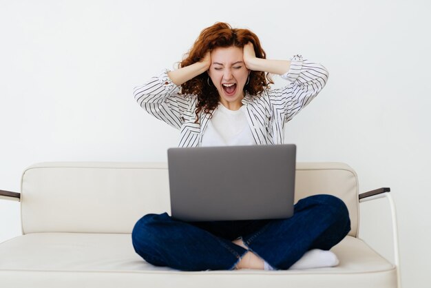 Wow Süße rothaarige Frau mit künstlichen Gliedmaßen, die auf dem grauen Sofa sitzt und auf den Laptop-Bildschirm schaut, während sie sich schockiert und überrascht fühlt Zeit zu Hause verbringen Konzept