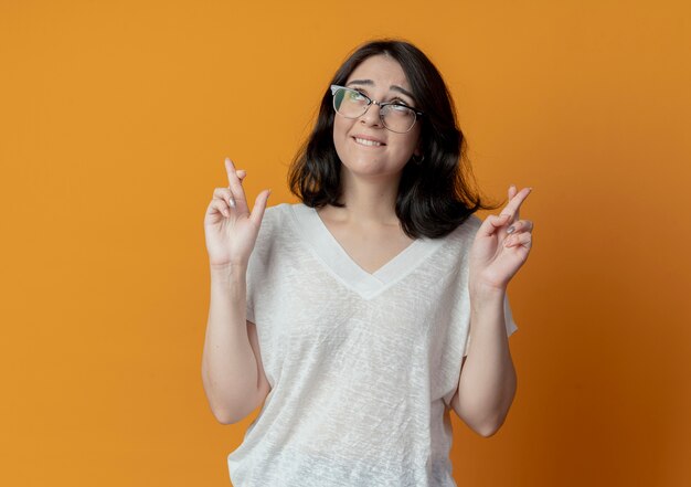 Wünschendes junges hübsches kaukasisches Mädchen, das eine Brille trägt, die nach oben schaut und gekreuzte Finger tut, lokalisiert auf orange Hintergrund mit Kopienraum