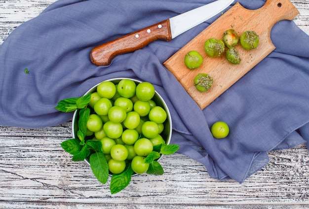 Würzige Greengages mit Blättern in einem Metalltopf und einem Holzschneidebrett mit Messerwinkelansicht auf einem grauen Holz und einem Picknicktuch