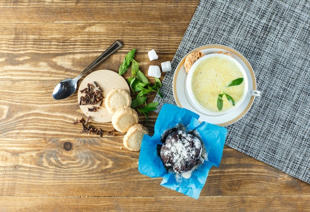 Würzige Milch mit Minze, Löffel, Zuckerwürfeln, Keksen, Nelken in einer Tasse auf Holztisch