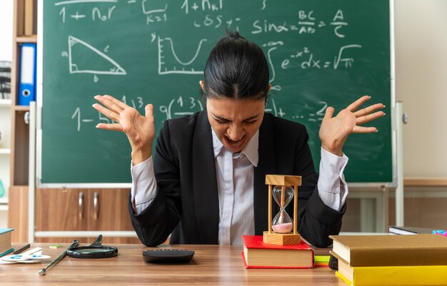 Wütend mit gesenktem Kopf sitzt die junge Lehrerin am Tisch mit Schulmaterial, das die Hände im Klassenzimmer ausbreitet