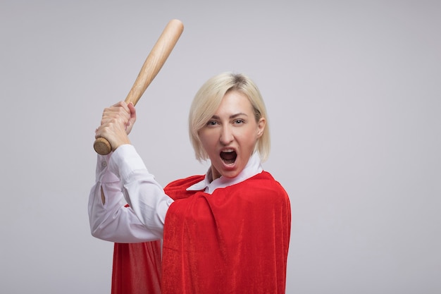 Wütende blonde Superheldin mittleren Alters in rotem Umhang, die Baseballschläger anhebt und auf die Vorderseite isoliert auf weißer Wand mit Kopierraum schaut