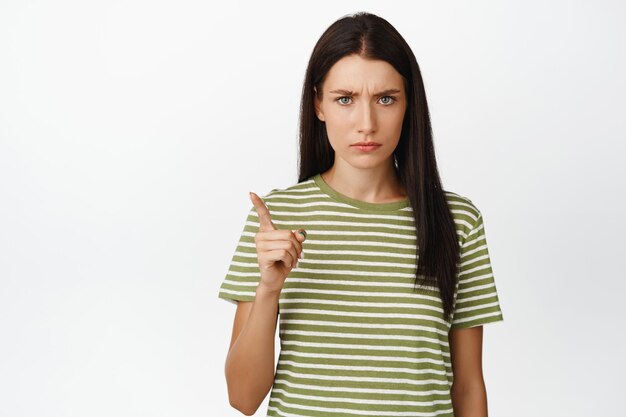 Wütende brünette Frau, die missbilligend den Finger schüttelt und schlechtes Benehmen schimpft, das auf weißem Hintergrund im T-Shirt steht