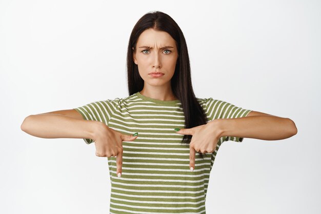 Wütende brünette Frau runzelt die Stirn und zeigt schmollend mit dem Finger auf etwas Schlechtes, das im T-Shirt vor weißem Hintergrund steht