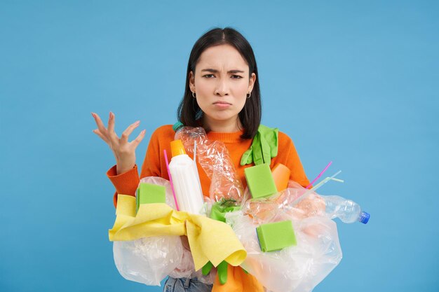 Wütende junge Frau ballt die Faust, hält unsortierten Plastikmüll und recycelt, sieht verärgert aus, blauer Hintergrund