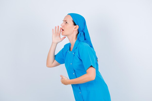 Wütende junge Krankenschwester schaut auf und schreit, indem sie die Hand in der Nähe des Mundes auf weißem Hintergrund hält