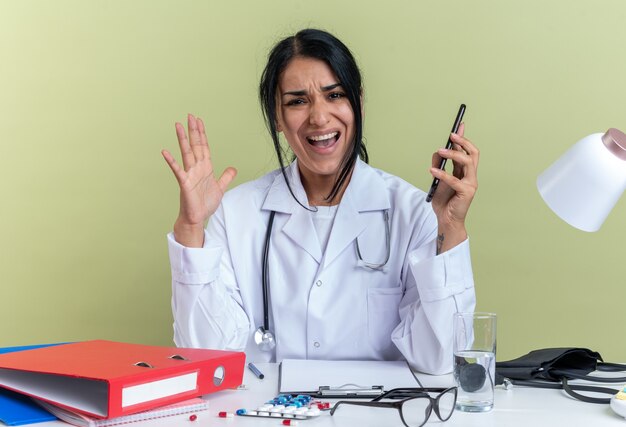 Wütende junge Ärztin, die medizinische Robe mit Stethoskop trägt, sitzt am Schreibtisch mit medizinischen Instrumenten, die das Telefon isoliert auf olivgrüner Wand halten