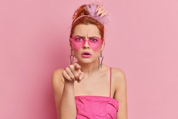 Wütende rothaarige junge frau trägt stylische sonnenbrille und kleid zeigt direkt mit verärgertem gesichtsausdruck etwas atemberaubendes isoliert über rosa wand Monochrome Aufnahme. Hallo du