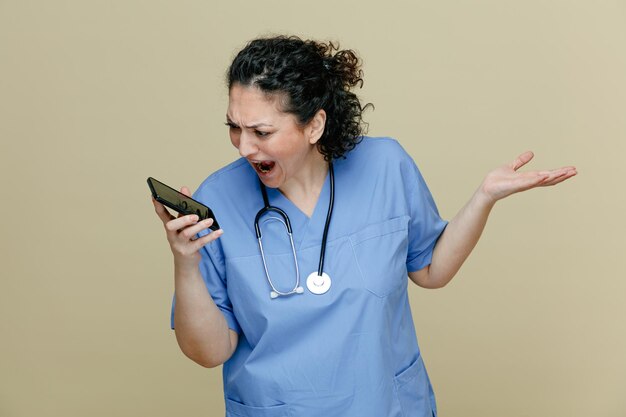 Wütende Ärztin mittleren Alters, die Uniform und Stethoskop um den Hals trägt und ein Handy hält, das es anschaut und eine leere Hand zeigt, die laut isoliert auf olivgrünem Hintergrund schreit