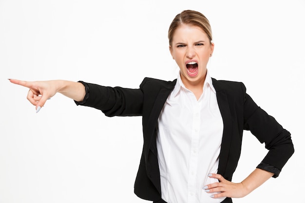 Wütende schreiende blonde Geschäftsfrau, die Arm auf Hüfte hält und über Weiß weg zeigt
