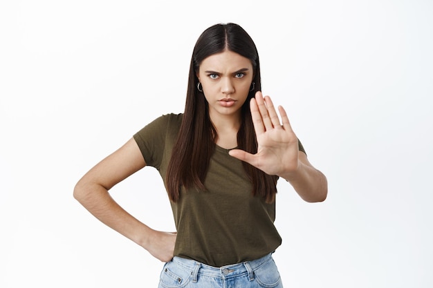 Wütende, stirnrunzelnde Frau streckt die Hand in Blockgeste aus, verbietet etwas Schlimmes und steht gegen weiße Wand