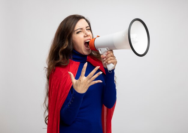 Wütende Superfrau mit rotem Umhang schreit in Lautsprecher, der auf weißer Wand isoliert ist