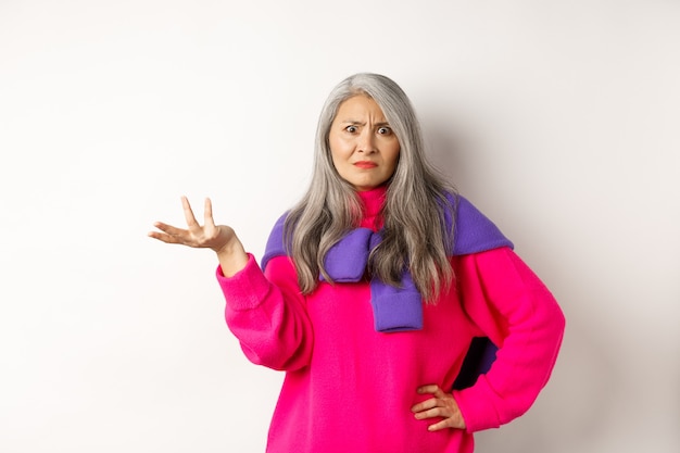 Wütende und verwirrte asiatische Seniorin breitete die Hand seitwärts aus und starrte verwirrt in die Kamera, stehend in rosa Pullover vor weißem Hintergrund