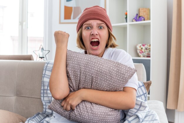 Wütende ungesunde junge Frau in warmem Hut mit Decke, die unwohl aussieht und an Erkältung und Grippe leidet, die Kissen mit geballter Faust schreit und mit aggressivem Ausdruck auf der Couch im hellen Wohnzimmer sitzt