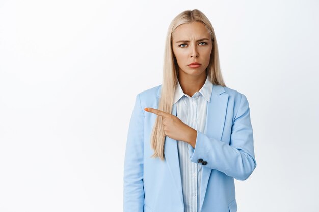 Wütende Verkäuferin, die nach links zeigt und missbilligend die Stirn runzelt, steht im Business-Anzug vor weißem Hintergrund