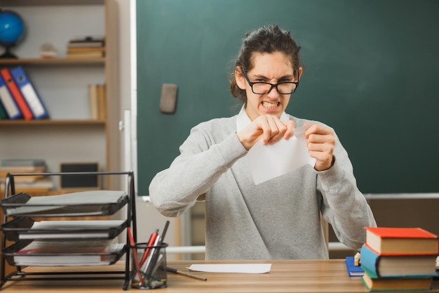 wütender Blick in die Kamera junger männlicher Lehrer mit Brille reißt Papier, das am Schreibtisch mit Schulwerkzeugen im Klassenzimmer sitzt