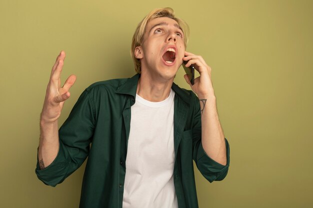 Wütender Blick nach oben junger blonder Kerl, der grünes T-Shirt trägt, spricht am Telefon, das Hand hebt