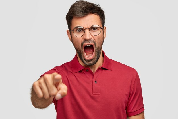 Wütender depressiver Mann mit dunklen Borsten, schreit jemanden wütend an, zeigt auf ein rotes T-Shirt, das über einer weißen Wand isoliert ist. Unrasierter Verrückter drückt Wut aus, schreit laut