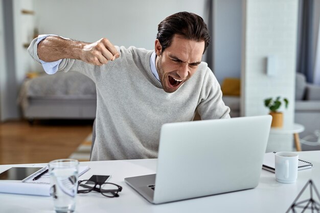 Wütender Geschäftsmann, der vor Frustration schreit und dabei ist, seinen Laptop zu schlagen, nachdem er schlechte Nachrichten gelesen hat, während er zu Hause arbeitet