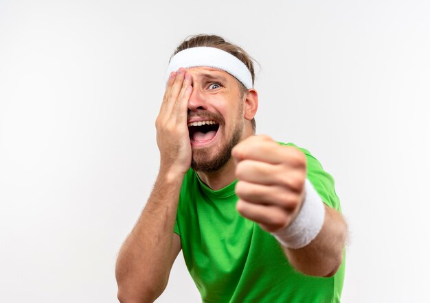 Wütender junger gutaussehender sportlicher Mann mit Stirnband und Armbändern, der die Hand auf das Gesicht legt und die Faust isoliert auf weißer Wand ausstreckt