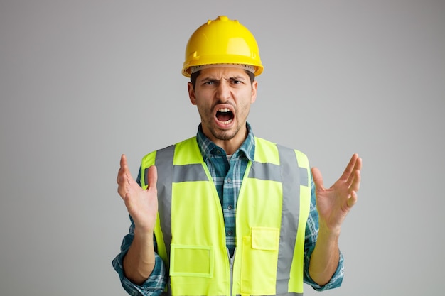 Wütender junger männlicher Ingenieur mit Schutzhelm und Uniform blickt in die Kamera und zeigt leere Hände, die isoliert auf weißem Hintergrund schreien