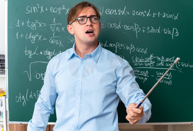 Wütender junger männlicher Lehrer mit Brille mit Zeiger, der die Lektion erklärt, die in der Nähe der Tafel mit mathematischen Formeln im Klassenzimmer steht