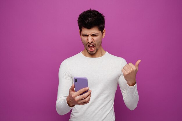 Wütender junger Mann, der ein Handy hält und anschaut, das auf die Seite zeigt und schreit, isoliert auf violettem Hintergrund