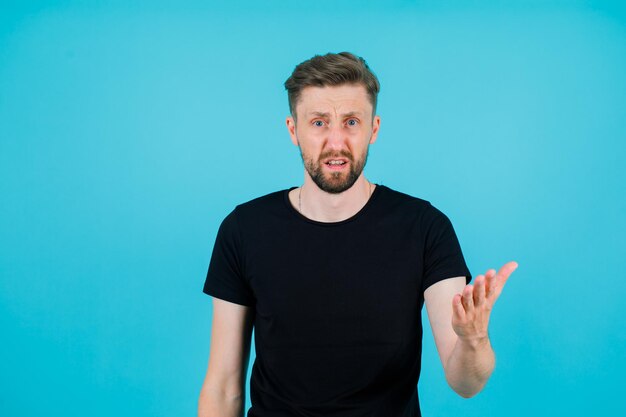 Wütender junger Mann schaut in die Kamera, indem er seine Hand auf blauem Hintergrund hochhält