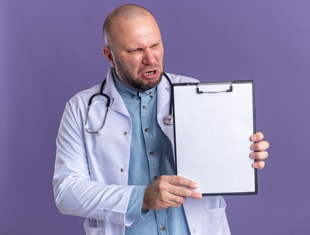 Wütender männlicher Arzt mittleren Alters, der ein medizinisches Gewand und ein Stethoskop trägt und die Zwischenablage zur Kamera zeigt, die sie isoliert auf lila Wand betrachtet