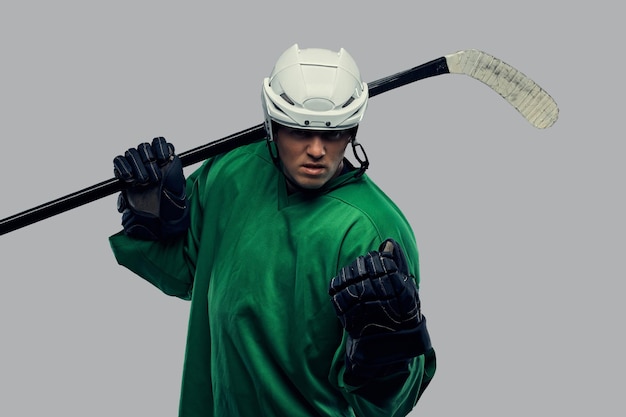 Wütender professioneller Hockeyspieler in grüner Sportbekleidung, der mit einem Hockeyschläger steht. Isoliert auf grauem Hintergrund.