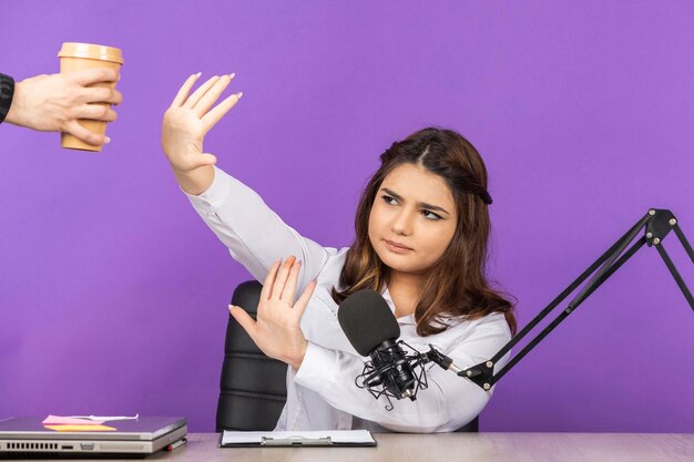 Wütender Radiomoderator lehnte Kaffee ab Junges Mädchen sitzt hinter dem Schreibtisch auf violettem Hintergrund Hochwertiges Foto
