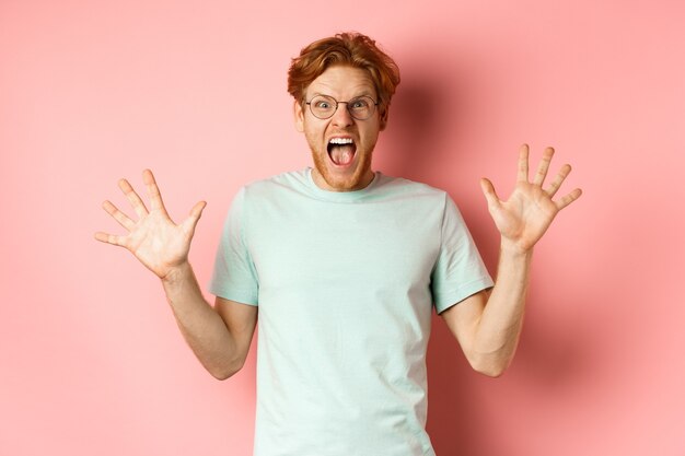 Wütender und unter Druck stehender junger Mann, der die Beherrschung verliert, die Hände zur Seite spreizt und mit wütendem Gesicht schreit und in Brille und T-Shirt vor rosa Hintergrund steht.