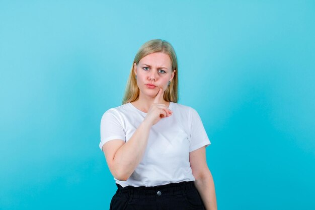 Wütendes blondes Mädchen schaut in die Kamera, indem es den Zeigefinger auf der Wange auf blauem Hintergrund hält