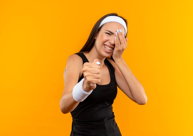 Wütendes junges hübsches sportliches Mädchen mit Stirnband und Armband, das ihre Faust ausstreckt und ihre Hand auf das Auge legt, das unter Schmerzen leidet, isoliert auf oranger Wand mit Kopierraum