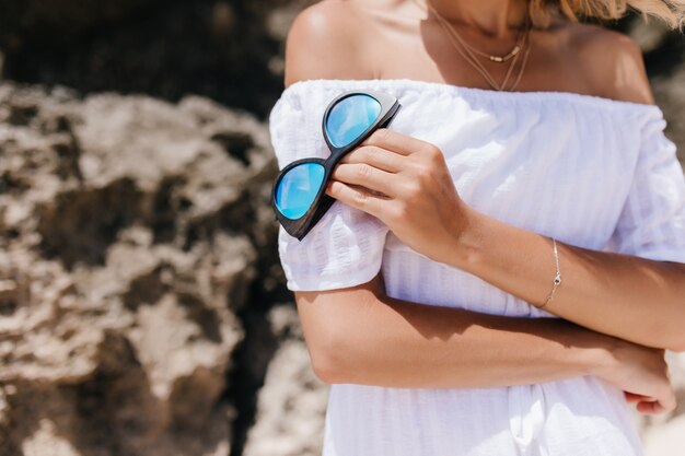 Wunderbare gebräunte Frau im eleganten Armband, das vor Felsen steht. Außenaufnahme der kaukasischen Frau im weißen Sommerkleid, das Gläser hält.