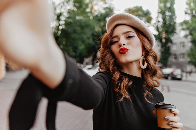 Wunderbare Ingwer Französisch Mädchen machen Selfie. Außenaufnahme der anmutigen lockigen Dame, die Tasse Cappuccino hält.