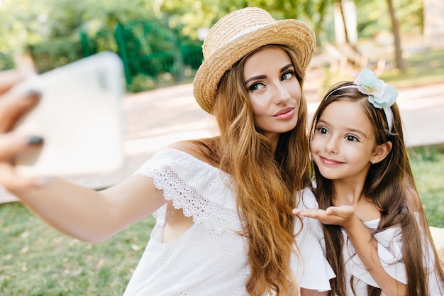 Wunderbare junge Dame mit hellem Make-up, die Smartphone hält und Selfie mit Tochter auf Naturhintergrund macht. Porträt eines dunkelhaarigen Mädchens, das einen Luftkuss für ein Foto mit Mutter sendet.