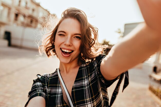 Wunderbare lockige Frau mit dunklen Augen, die spielerisch auf der Straße posieren. Foto im Freien von inspirierter junger Dame, die selfie macht.