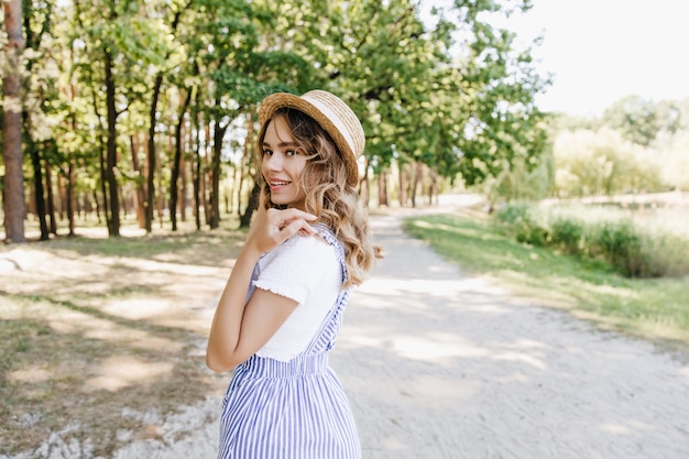 Wunderbares blondes Mädchen, das im Sommerpark geht und lächelt. Foto im Freien von entzückender Dame im Strohhut, der mit Vergnügen auf Natur aufwirft.