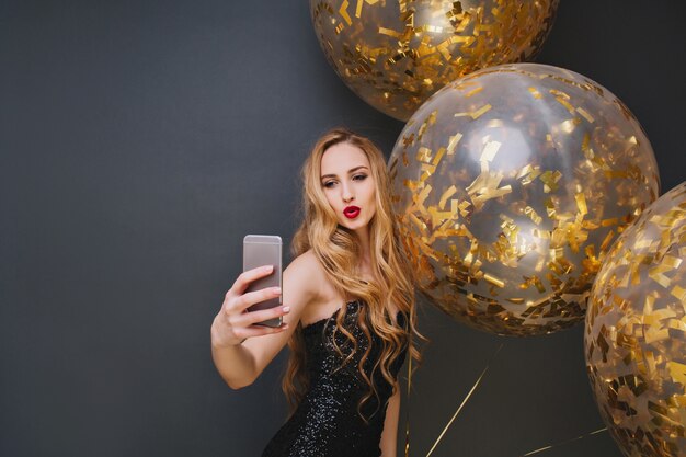 Wunderbares europäisches Mädchen, das selfie mit küssendem Gesichtsausdruck macht. Prächtige junge Frau mit langen Haaren, die Geburtstagsfeier mit großen Luftballons genießt.