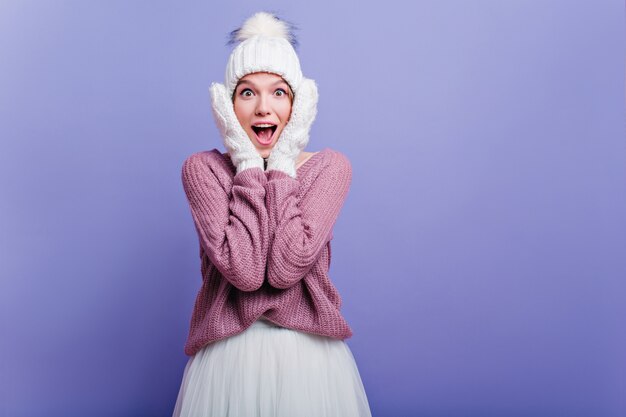 Wunderbares Mädchen in den weißen gestrickten Handschuhen, die Glück mit lila Wand ausdrücken. Innenfoto des hübschen jungen weiblichen Modells im Hut, der mit Erstaunen aufwirft.