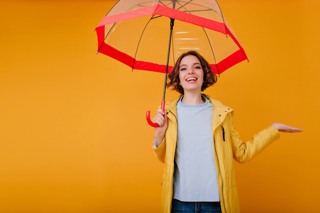 Wunderbares Mädchen in guter Laune, das lacht, während es mit rotem Regenschirm aufwirft. Innenfoto der trendigen kaukasischen Dame mit funkelndem Make-up, das Fotoshooting mit Sonnenschirm genießt.