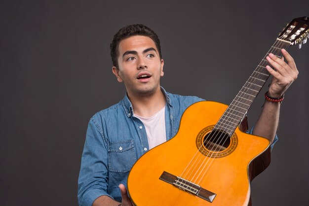 Wundernder Mann, der die Gitarre auf schwarzem Hintergrund hält. Hochwertiges Foto